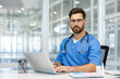 © Liubomir - Doctor in blue scrubs using laptop in office setting, highlighting healthcare, technology, online, and management. Glass windows provide bright, modern workspace ambiance.