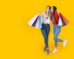 Shopping offer, smiling two young caucasian full body length view woman posing with shopper bags holding hands over yellow color background. Looking copy space. Happy customer concept idea photo.