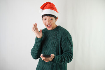 Excited young asian man wearing santa clause hat and green long sleeved sweater is using smartphone, isolated over white background. Concept for Christmas Holiday and New Year Party