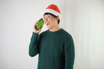 Excited young asian man wearing santa clause hat and green long sleeved sweater is calling someone using smartphone, isolated over white background. Concept for Christmas Holiday and New Year Party