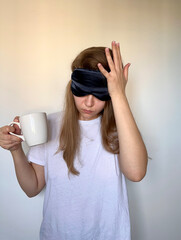 a woman wearing a black sleep mask, holds her head and a coffee mug, appearing tired, sleepy or havi