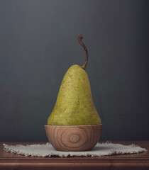 A pear in a wooden bowl standing on a linen napkin on a carved table on a dark green background. 