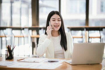 Beautiful Asian female accountant working with smartphone and laptop at her desk analyzing business reports and documents. Sending messages