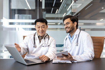 Two smiling doctors in white coats discussing medical information using a laptop and phone in a modern office. Concept of teamwork, communication, and technology in healthcare.