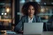 © N Joy Art  - Professional African American businesswoman with curly hair sitting at a desk in a modern office, using a laptop. Soft lighting, focused expression, wearing a gray blazer, late-night working