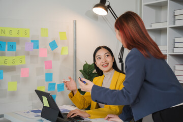 Brainstorming Success: Two businesswomen collaborate on a project, with sticky notes covering the wall behind them, in a modern office setting.