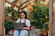 © sashafolly - Happy asian lady sitting with portable computer by doors of greenhouse and surfing information about plants
