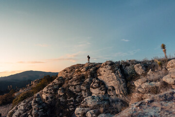 A lo lejos un Joven fotógrafo y explorador latino sosteniendo su cámara, fotografiando el horizonte, de pie sobre rocas en un paisaje de montañas.