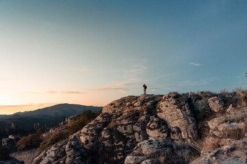 A lo lejos un Joven fotógrafo y explorador latino sosteniendo su cámara, fotografiando el horizonte, de pie sobre rocas en un paisaje de montañas.