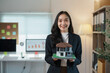 ©  NCST Studio - Smiling real estate agent holds a model house in her office, assisting clients with property investments, sales, rentals, and purchases in a modern workspace