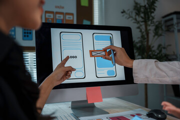 Two web designers are working on a user interface for a mobile application, placing elements on a wireframe displayed on a computer screen