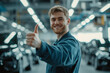 © ALL YOU NEED studio - attractive car mechanic smiling and holding up his right hand with the thumb pointing up . He is wearing blue overalls.