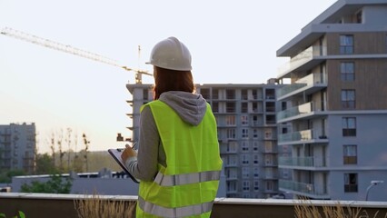 Construction female engineer taking notes and writing on clipboard while inspecting a building site at sunset. Industry development concept