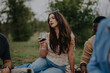 © qunica.com - A young woman holds a glass of red wine, surrounded by friends at an outdoor picnic. The setting is peaceful, highlighting relaxation and friendship in a lush, green environment.