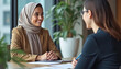 © LuisAlfonso - Chatting with a smiling European colleague at the meeting table is a contented young Arab businesswoman wearing a headscarf. Speaking about collaboration and a project, two young, varied businesswomen