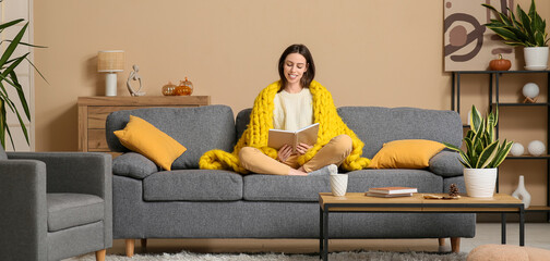 Beautiful young woman with warm yellow plaid reading book while sitting on sofa in living room