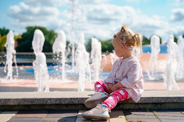 Caucasian cute little child toddler girl walks by the fountains in summer.