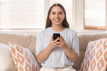 Poster - Smiling young woman using mobile phone on sofa at home