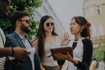 A multicultural business group gathers outside, discussing and brainstorming project ideas and strategies for growth. The team collaborates to analyze and strategize in a bustling urban environment.