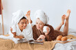 © Prostock-studio - Positive beautiful multiracial girlfriends in bathrobes and towels on heads drinking coffee with croissants, reading magazines together while laying on bed at home