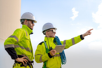 two engineers in high visibility gear and hard hats observe a wind turbine and communicate via radio