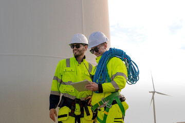 two engineers in high visibility gear stand near a wind turbine, reviewing a clipboard and preparing