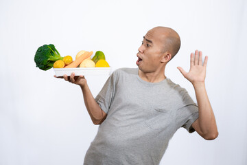 happy and smiling bald Asian man standing with a tray full of healthy and nutritious fresh vegetables with finger pointing, concept healthy lifestyle and eating nutritious vegetables, isolated white.
