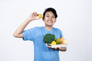 Young excited Indonesian man standing carrying tray of healthy vegetables and fruits with his hand pointing, healthy lifestyle concept, healthy food campaign, isolated on white background.