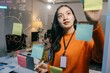 © Parichat - Young businesswoman is using adhesive notes to plan a project on a glass wall in an office. She is wearing an orange sweater and has a lanyard around her neck