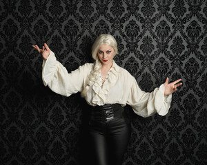 Canvas Print - close up portrait of beautiful blonde female model, wearing vintage gothic fantasy ruffle blouse white shirt, leather pants and long blonde plaited hair.  moody black brocade wall paper background