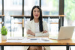 © Nittaya - Confident Businesswoman:  A young, confident Asian businesswoman sits at her desk, arms crossed, radiating success and determination.