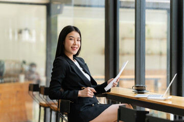 Confident Businesswoman: A young Asian businesswoman exudes confidence and professionalism, working on documents in a modern cafe setting. Her smile radiates success and accomplishment.