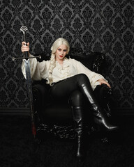 Poster -  Full length portrait of blonde female model wearing vintage gothic fantasy blouse, leather pants. elegant sitting pose black ornate throne chair, holding sword weapon, brocade wallpaper backdrop