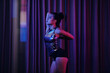 © DragonImages - Asian woman in metallic dance outfit standing by pole in dimly lit room, reflecting on her practice. Curtains create backdrop for serene and focused moment
