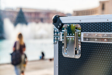 The metal-cornered case rests near a tranquil waterfront where people are gathered, enjoying leisure time under the bright sun in the background.