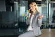 © Liubomir - Mature businesswoman in gray suit having phone conversation while holding glasses, sitting on desk confidently. Shows leadership, communication, professionalism in modern office environment.
