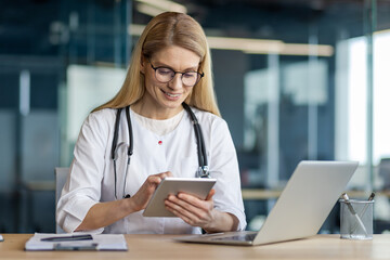 Wall Mural - Smiling female doctor using tablet in office. Wearing white coat, stethoscope, glasses, working on laptop. Represents healthcare, technology, professionalism, medical practice.