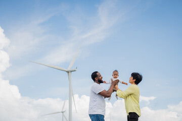 A family of three, a man and two women, are holding a baby. The sky is blue with some clouds. electrical, engineer, environment, energy, clean, sustainable, wind turbine, nature