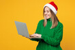 © ViDi Studio - Merry fun young IT woman wear green warm cozy sweater Santa hat posing hold use work on laptop pc computer isolated on plain yellow background. Happy New Year, celebration Christmas holiday concept.
