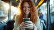 © familymedia - A young woman with vibrant red hair beams a smile while using her smartphone on a bus, depicting joy, connectivity, and the blend of public transit with technology.