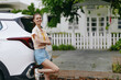 © SHOTPRIME STUDIO - Young woman smiling beside a white SUV, casual summer attire, sunny outdoor setting with greenery and a charming house in the background Relaxed and cheerful mood