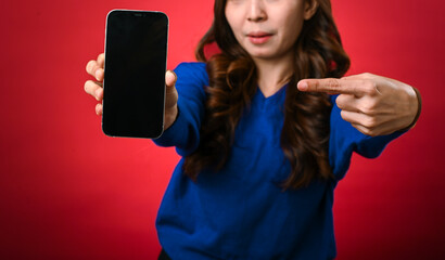 An Asian woman in a blue sweater is smiling and pointing at a smartphone she's holding forward, displaying its black screen. The background is a bold red, creating a striking contrast