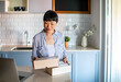© Marko Geber - Smiling Asian woman opening package at home with laptop