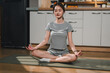 © kenchiro168 - Meditating woman in gray outfit sitting cross legged on yoga mat, practicing mindfulness in modern kitchen. serene expression reflects tranquility and focus