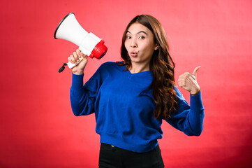 An Asian woman wearing a blue sweater is holding a red and white megaphone while speaking into it. She appears to be shouting or making an announcement. The background is solid red