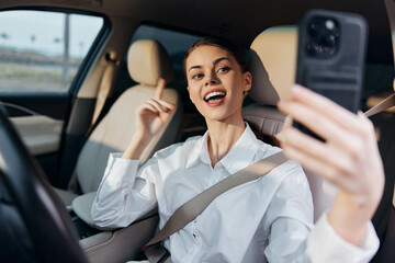 Woman taking a selfie in car, sitting in driver's seat with one hand on steering wheel, using cell phone, casual outfit, inside vehicle, daytime