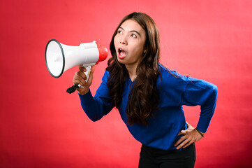 An Asian woman wearing a blue sweater is holding a red and white megaphone while speaking into it. She appears to be shouting or making an announcement. The background is solid red