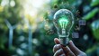 © Amornrat - Hand holding a glowing light bulb with green energy symbols, representing sustainable power and innovation in a natural setting.
