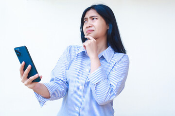 Wall Mural - thoughtful asian woman using smartphone with hands on chin thinking problem wearing blue striped casual shirt standing over isolated white background, look away