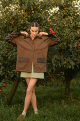 A beautiful girl in a brown autumn jacket in an apple orchard. A fashionable girl at a fashion show in an apple orchard